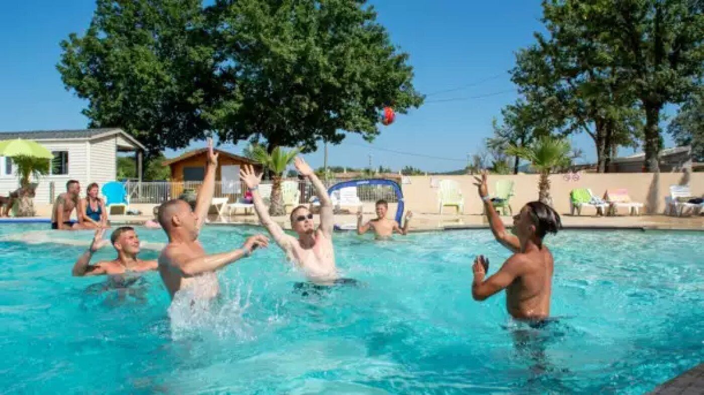 Una piscina con gente jugando, rodeada de árboles y un edificio.