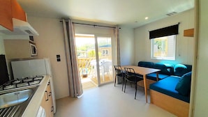 A compact kitchen with a gas stove, a microwave, and a small refrigerator. There is a dining area with a table and chairs, and a blue sofa. A sliding glass door leads to an outdoor area with a wooden deck and chairs.