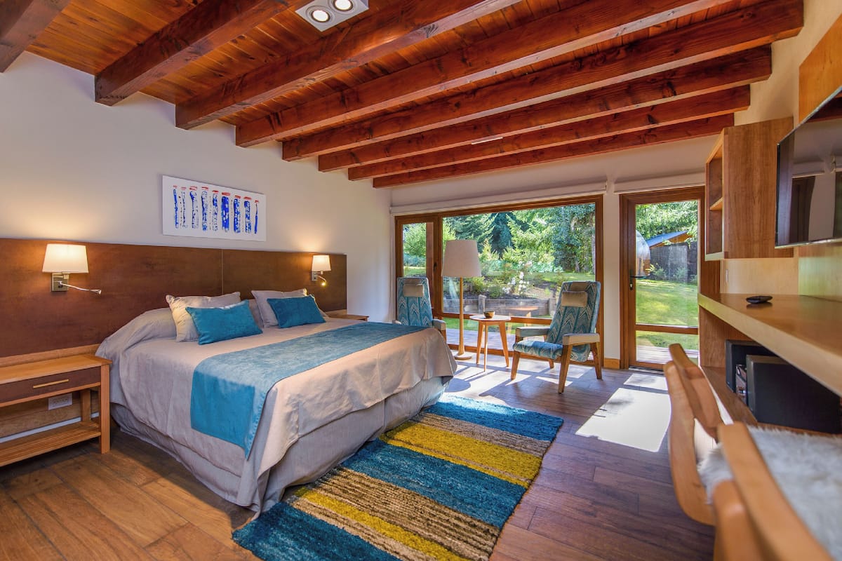 Une chambre avec un grand lit, des poutres apparentes en bois au plafond, un bureau et une vue sur l’extérieur.
