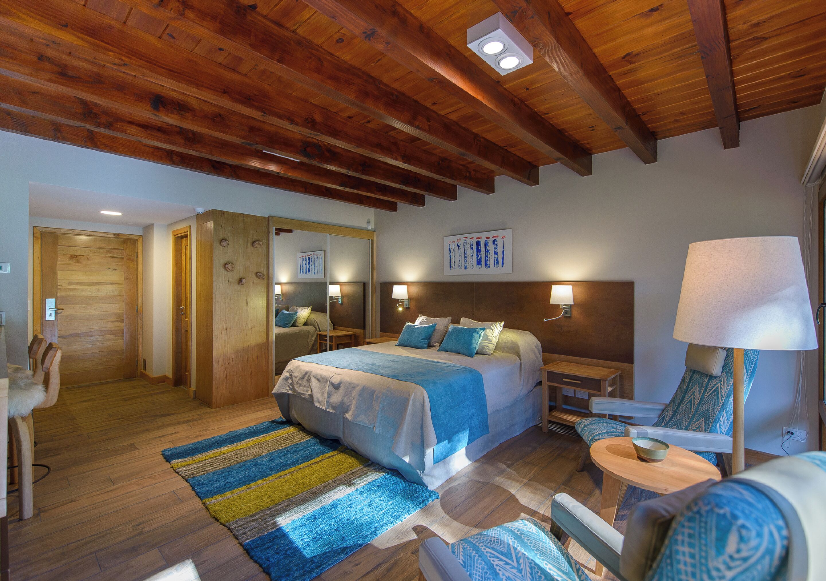Ein Hotelzimmer mit Holzboden, einem Bett mit blauer Bettwäsche, einem Nachttisch mit Lampe, einem Sessel mit gemustertem Kissen und einem blau-gelb gestreiften Teppich.