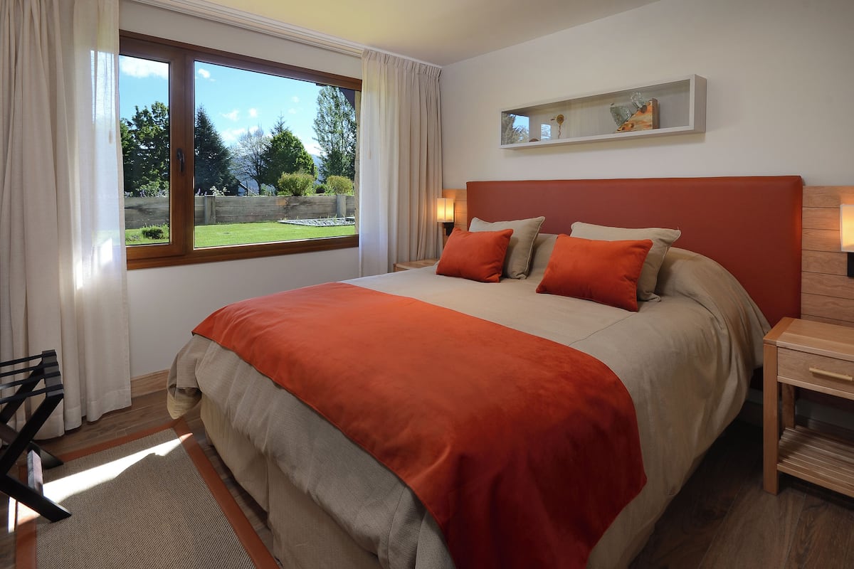 Une chambre avec un grand lit, une table de chevet, une commode en bois, une fenêtre avec des rideaux et une vue sur l’extérieur.