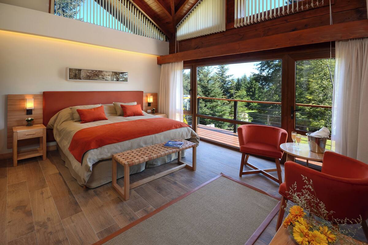 Une chambre avec un grand lit, un banc, une table avec un vase, et une vue sur les arbres à travers de grandes fenêtres.
