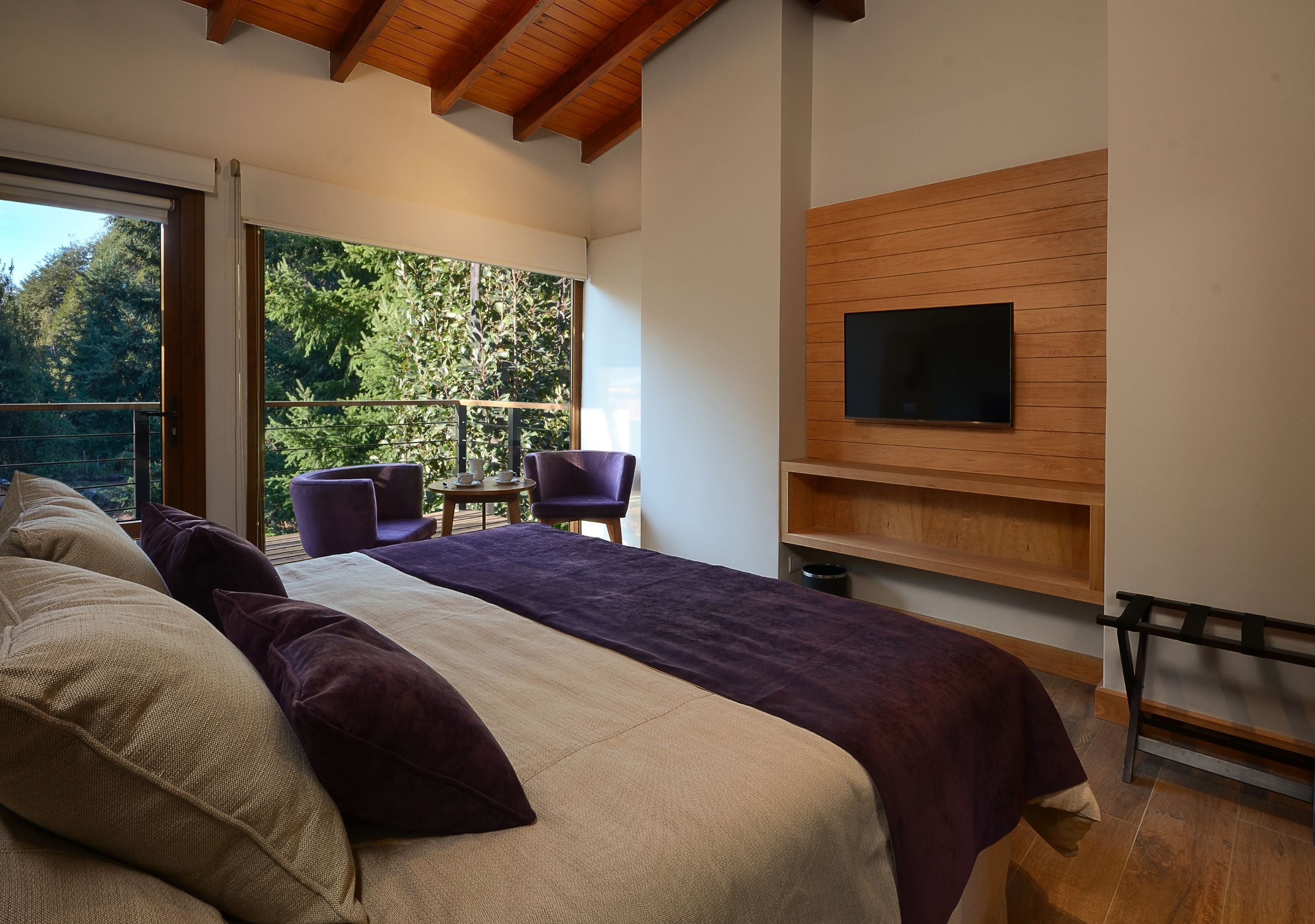 A bedroom with a large bed, a TV mounted on the wall, and a balcony with chairs.