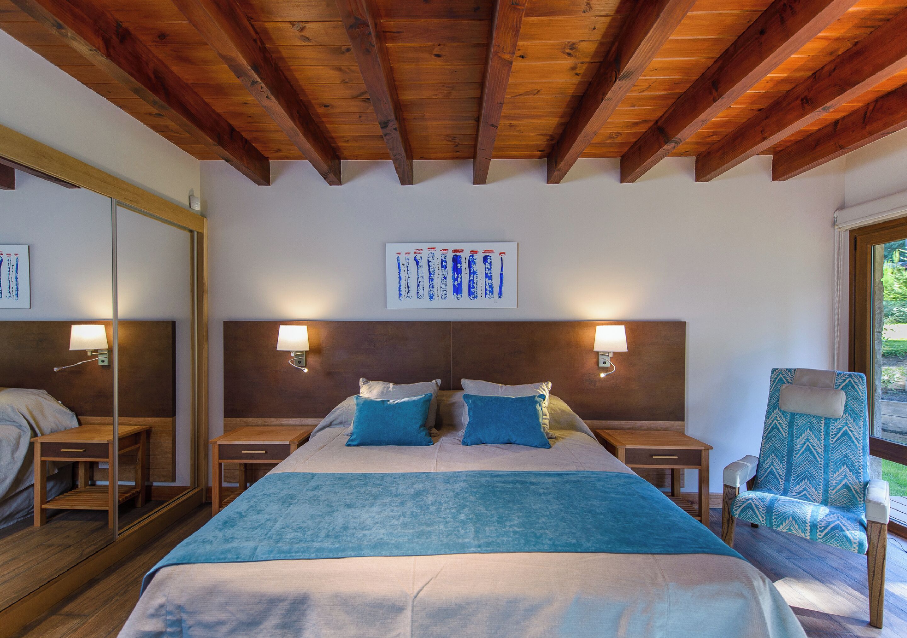 Hotelkamer met een houten plafond, een groot bed met blauw beddengoed, twee nachtkastjes met lampen, een stoel en een raam met uitzicht op groen.