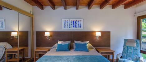 Hotelkamer met een houten plafond, een groot bed met blauw beddengoed, twee nachtkastjes met lampen, een stoel en een raam met uitzicht op groen.