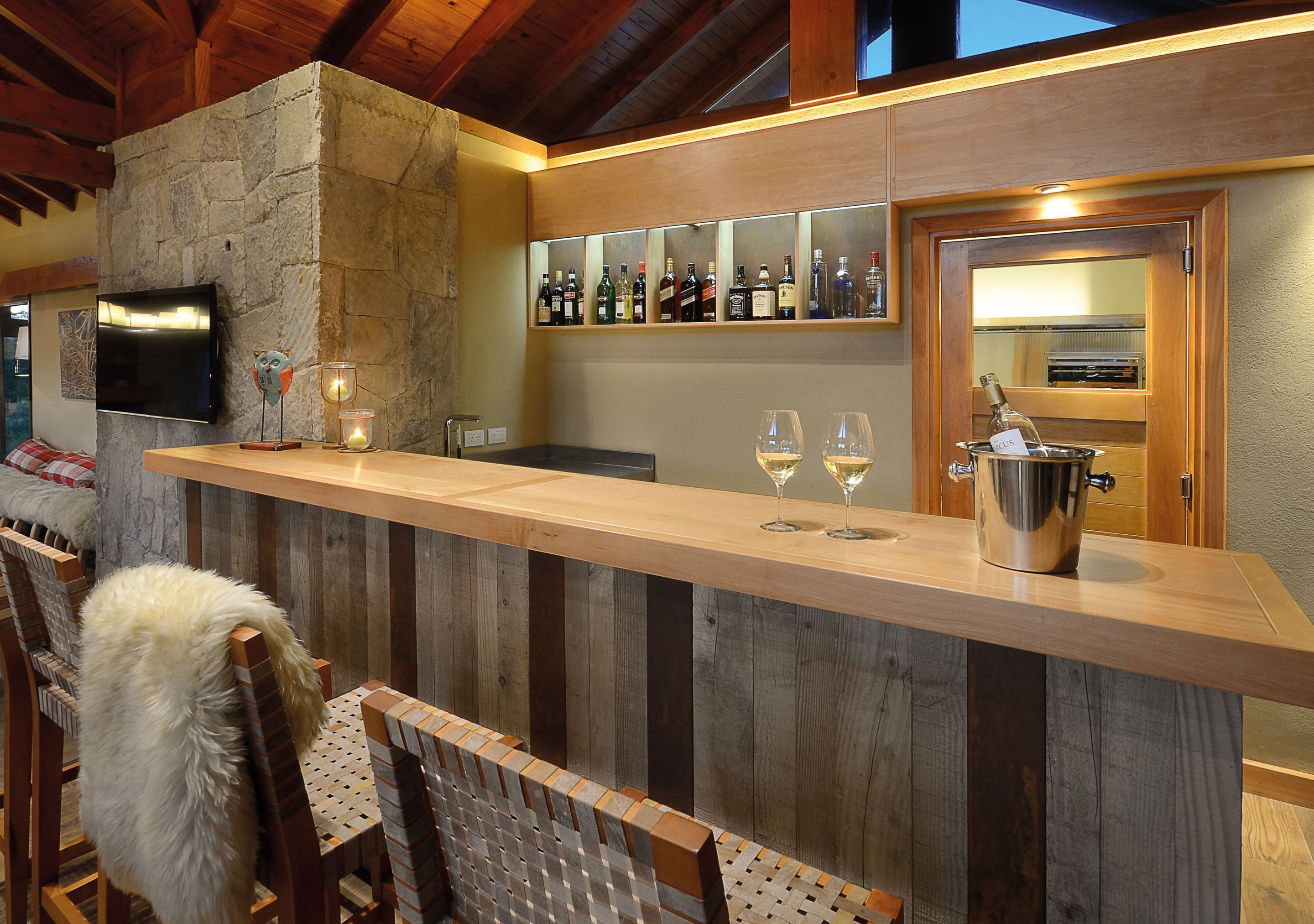 A bar area with a stone wall, wooden ceiling, and a shelf with bottles. There are two glasses on the counter and a candle.