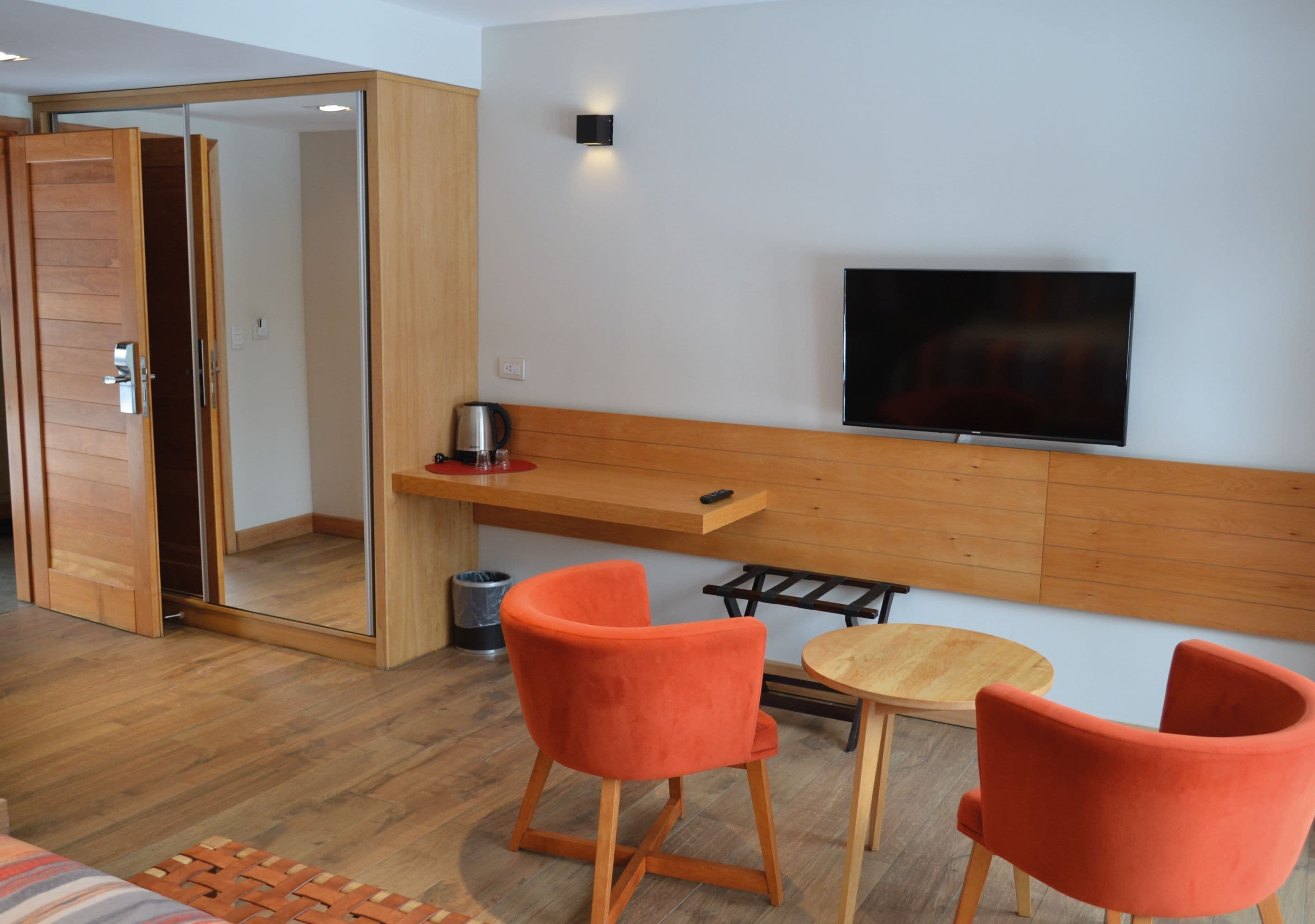 A modern hotel room with a wooden floor, a TV mounted on the wall, two orange chairs, a small wooden table, and a sliding door closet.