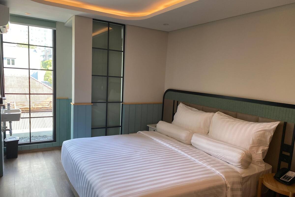 A hotel room with a large bed, a bedside table with a phone, and a window with a view of buildings outside.