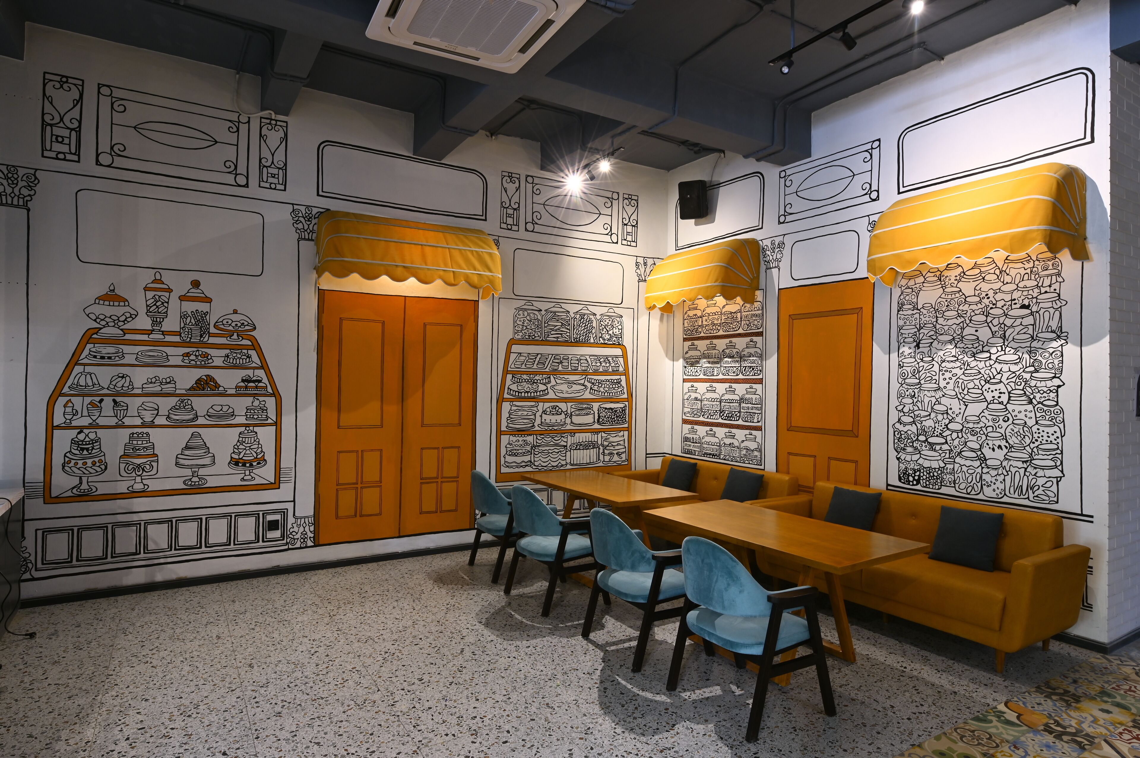 A room with a long wooden table, blue chairs, and a sofa. There are wall decorations with black and white illustrations of food and kitchen items.