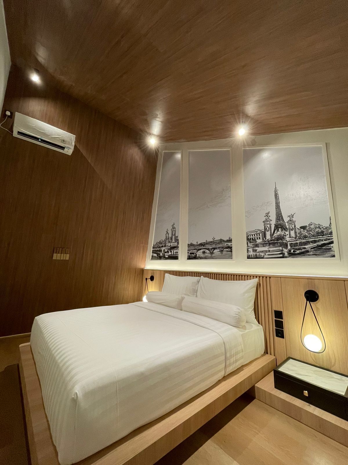 Una camera d'albergo moderna con soffitto in legno, un letto con lenzuola bianche, un condizionatore d'aria a parete e un'ampia finestra con vista sulla città.