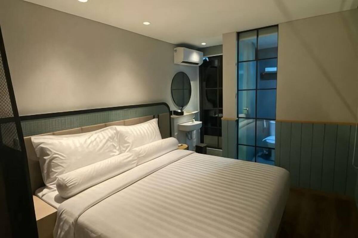 A hotel room with a bed, a bedside table, a mirror, a window, and a bathroom visible through the glass wall.