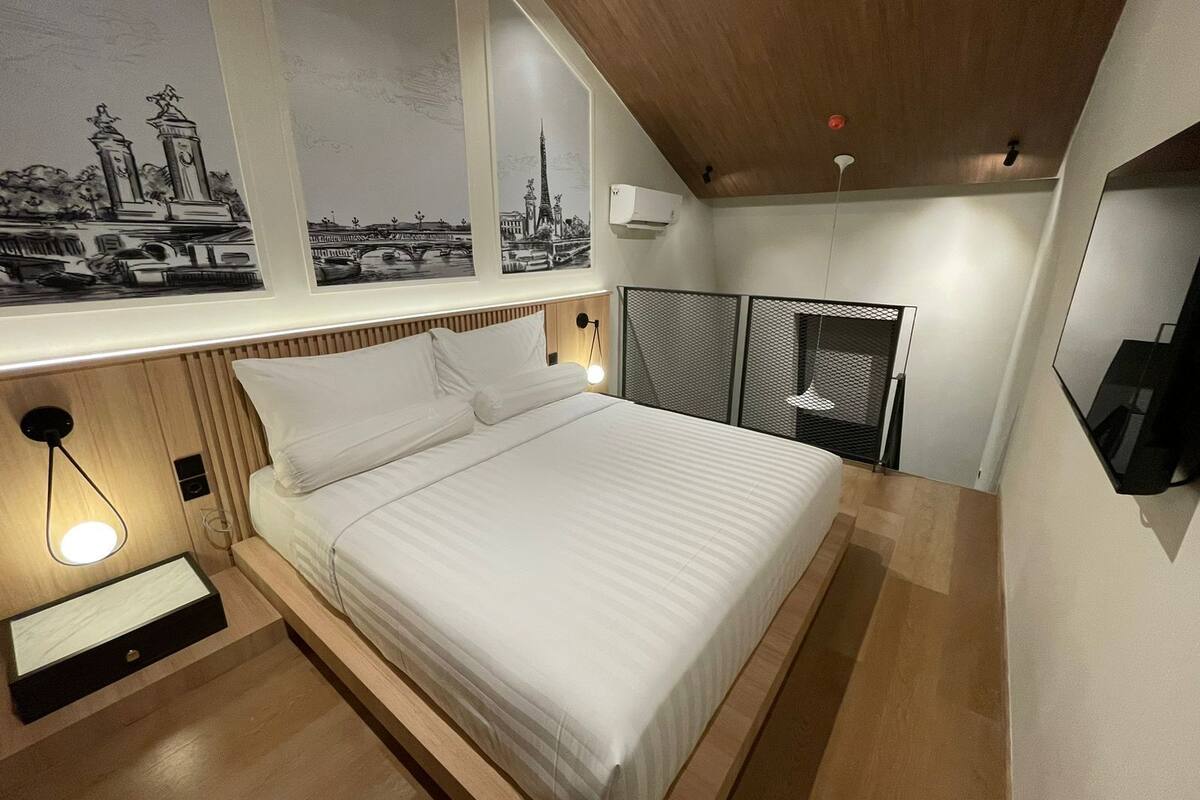 A modern bedroom with a large bed, a window with cityscape artwork, a wall-mounted TV, and a ceiling light.