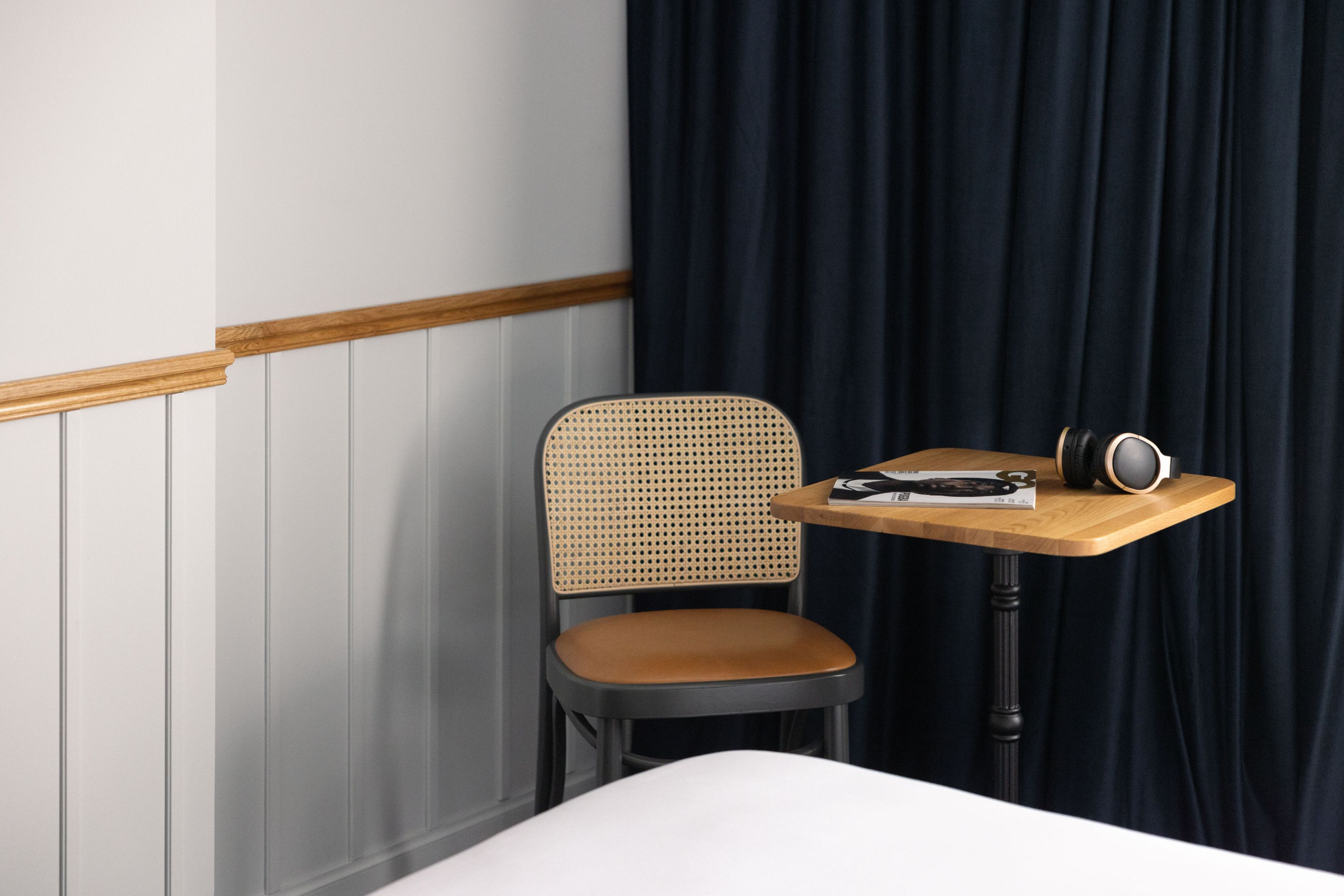Une chambre d’hôtel avec un lit, une table de chevet en bois, une chaise avec un dossier en osier et un rideau foncé.