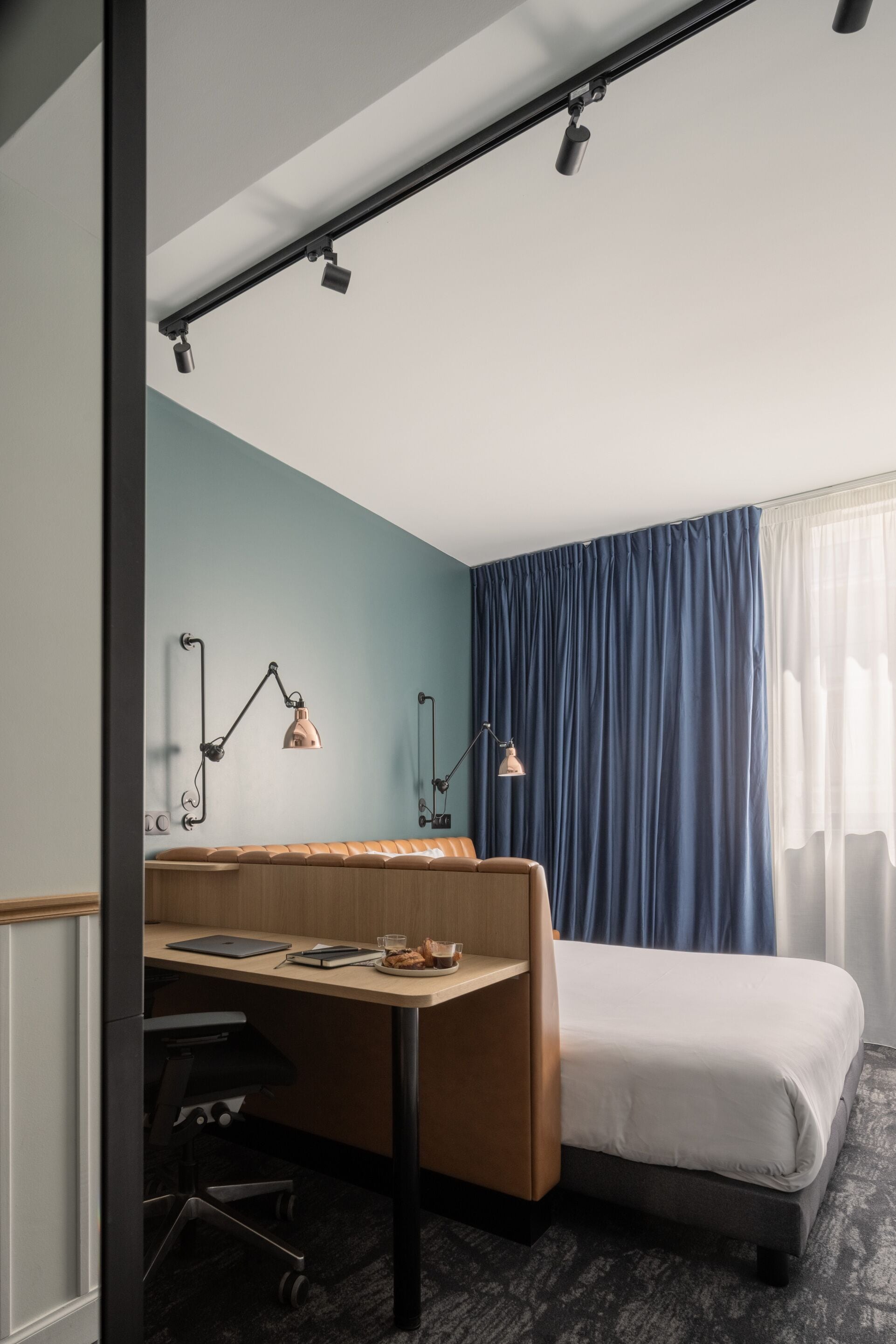 A hotel room with a desk, two lamps, a bed, and a view of the outdoors through a window with curtains.