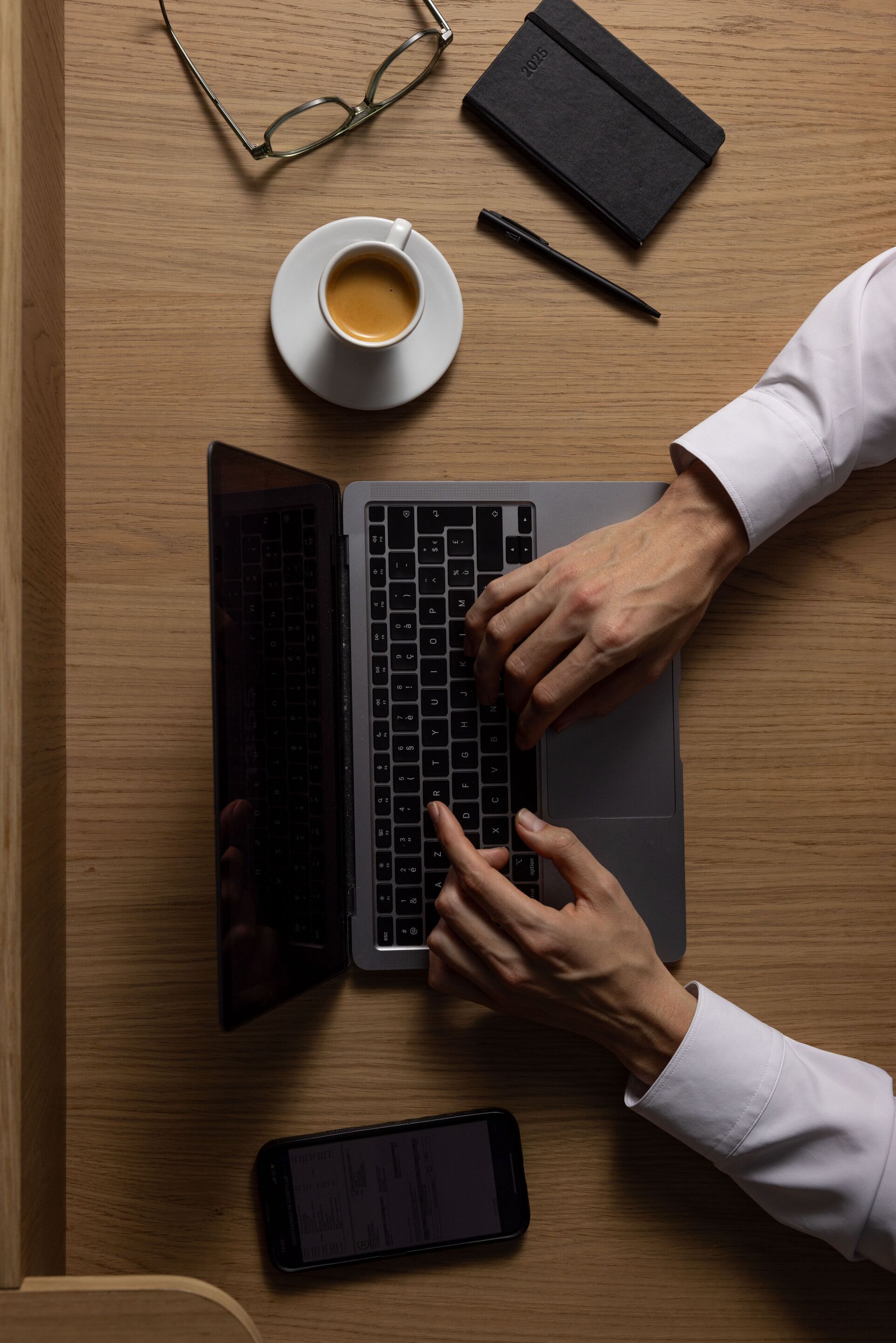 Un ordinateur portable sur une table en bois, avec un smartphone, des lunettes et un carnet.