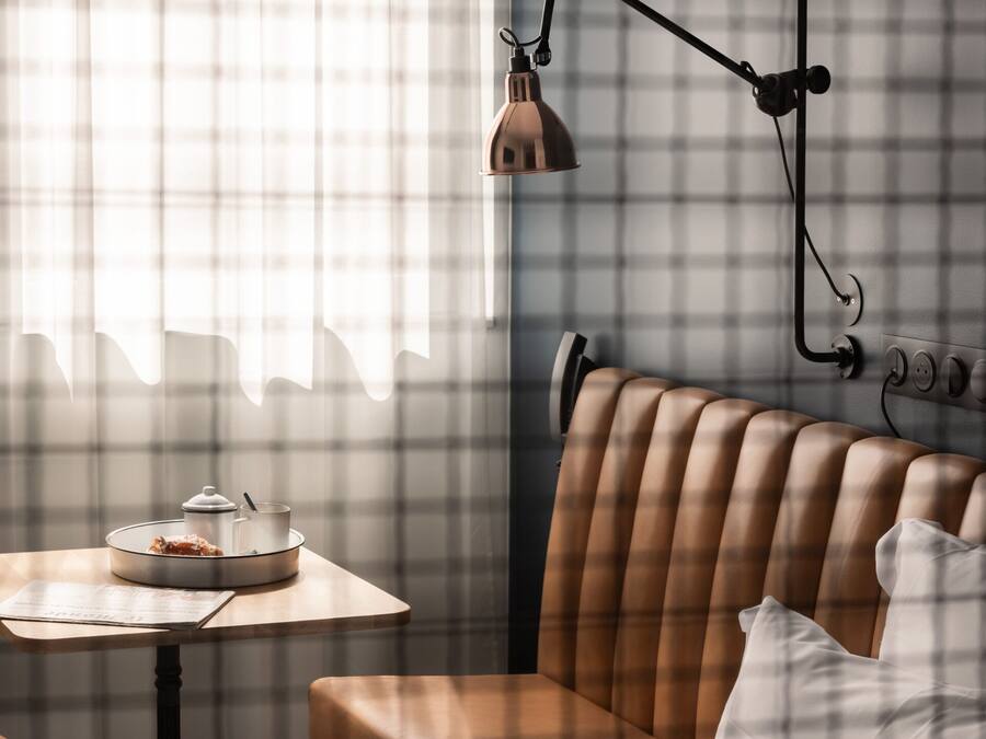 Una habitación acogedora con una pared a cuadros, un banco de cuero marrón, una mesa con lámpara y una ventana con cortinas traslúcidas.