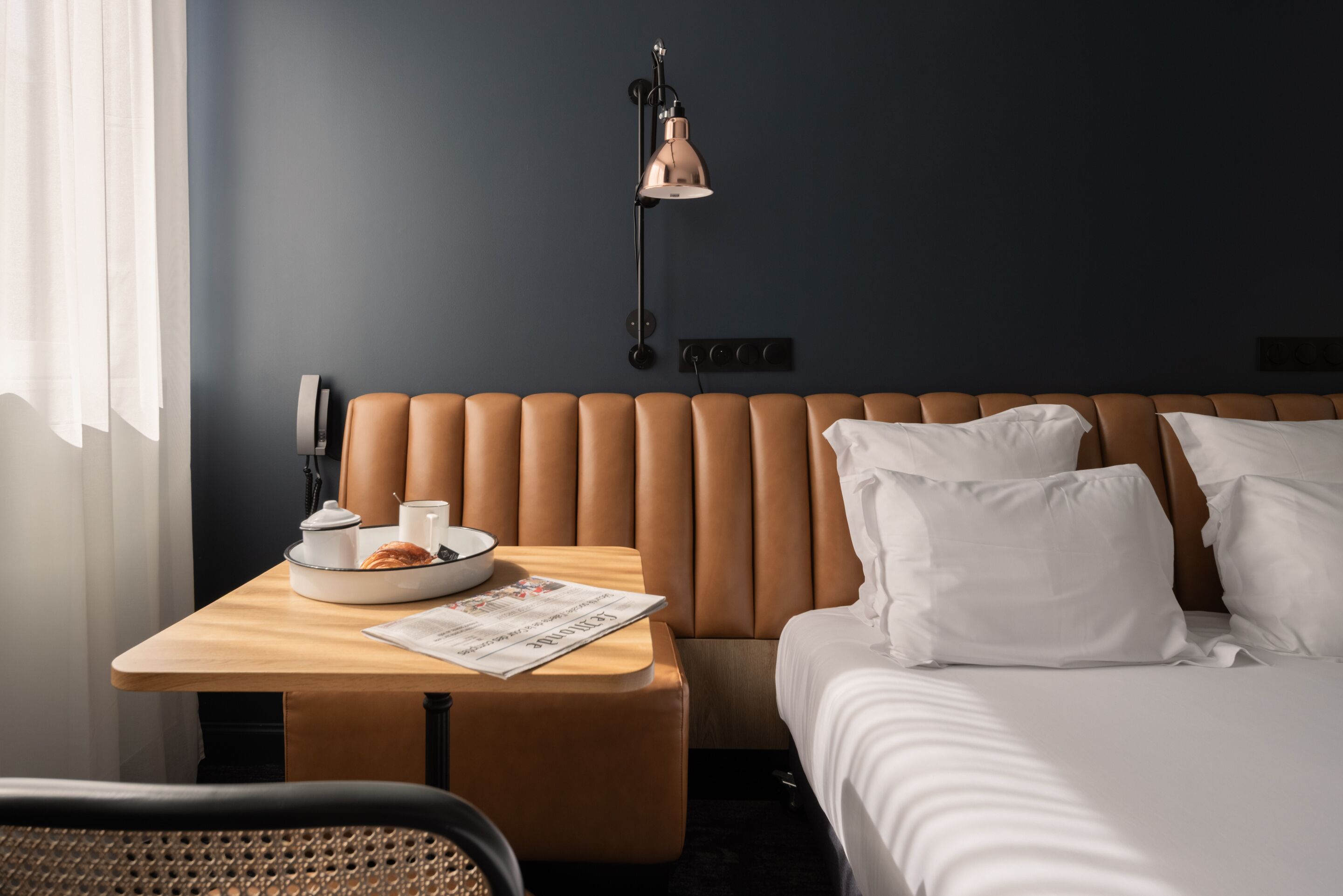 Ein Hotelzimmer mit Bett, Nachttisch mit Zeitung, Lampe und Frühstückswagen.