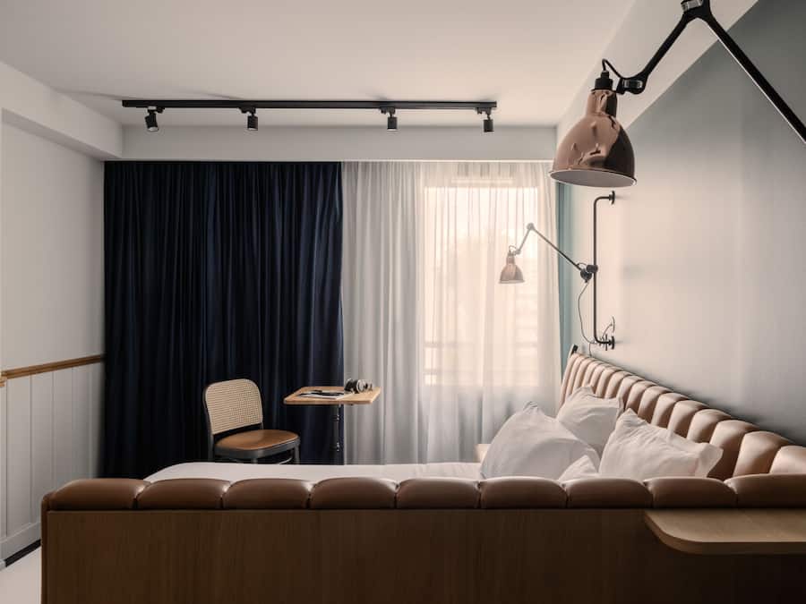 A modern hotel room with a curved bed, a small desk, a chair, and a large window with curtains.