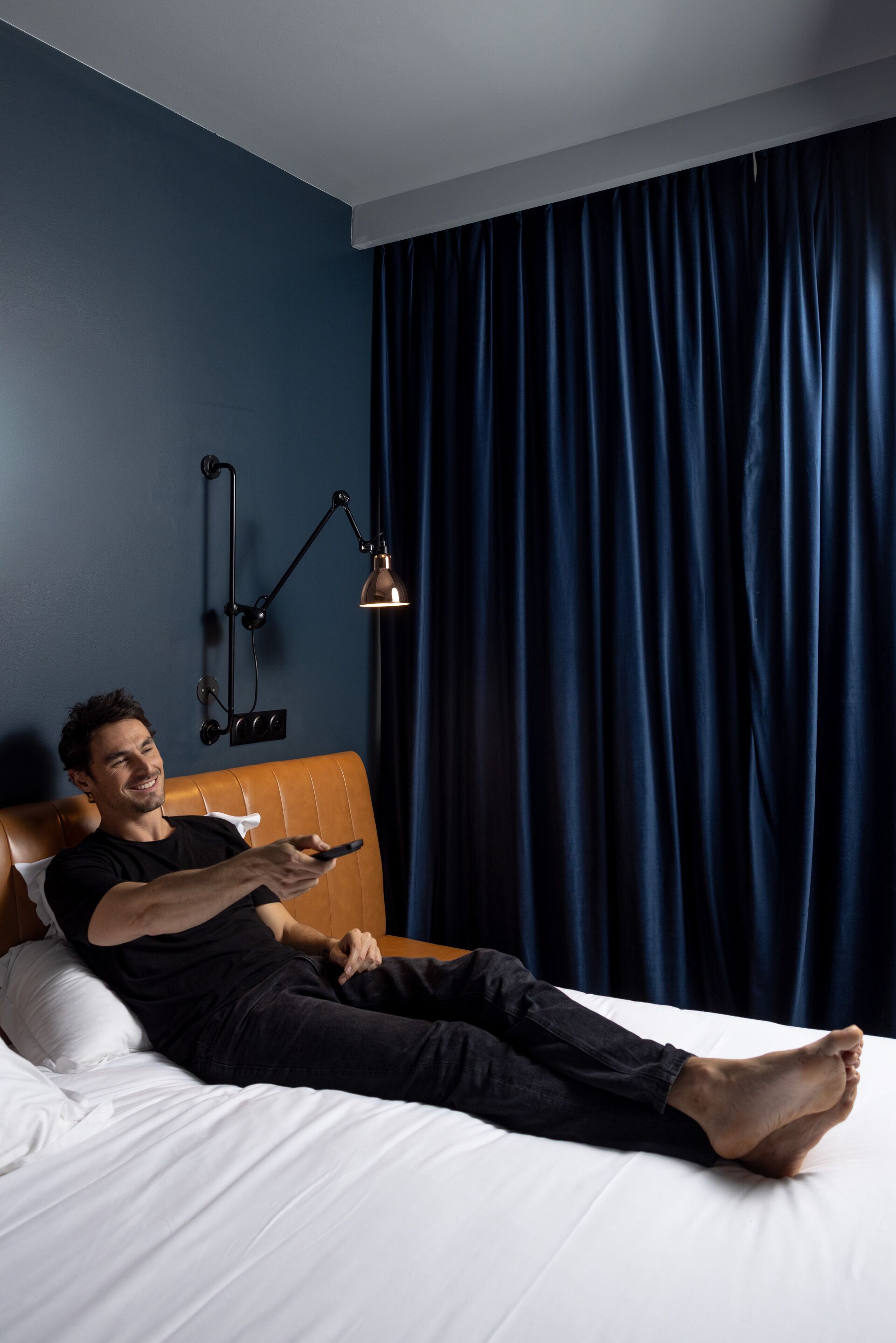 Una persona recostada en una cama en una habitación con pared azul oscuro, cabecera de madera y lámpara de escritorio negra.