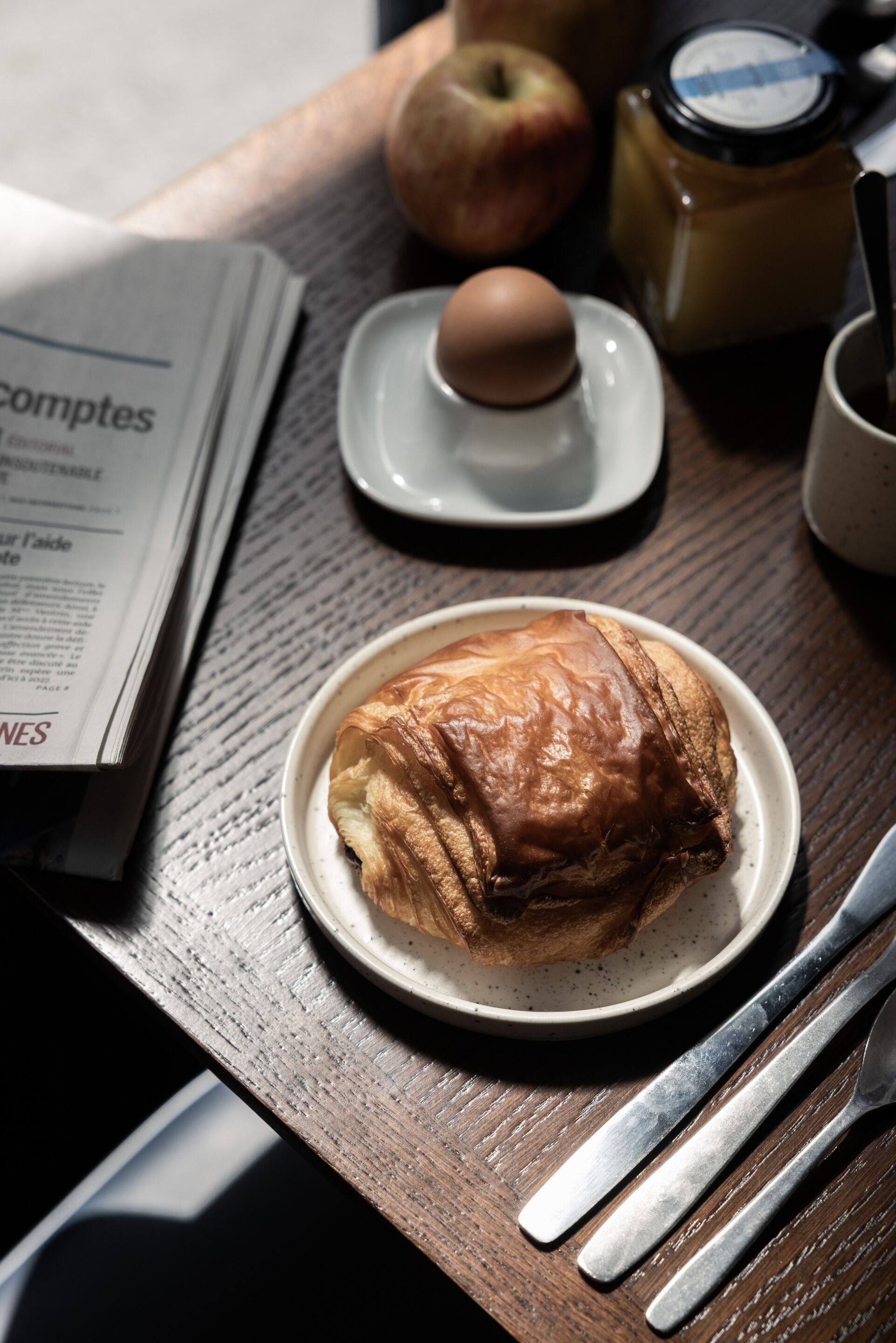 Une table en bois avec un croissant, une assiette contenant un œuf, un pot de confiture et un journal.