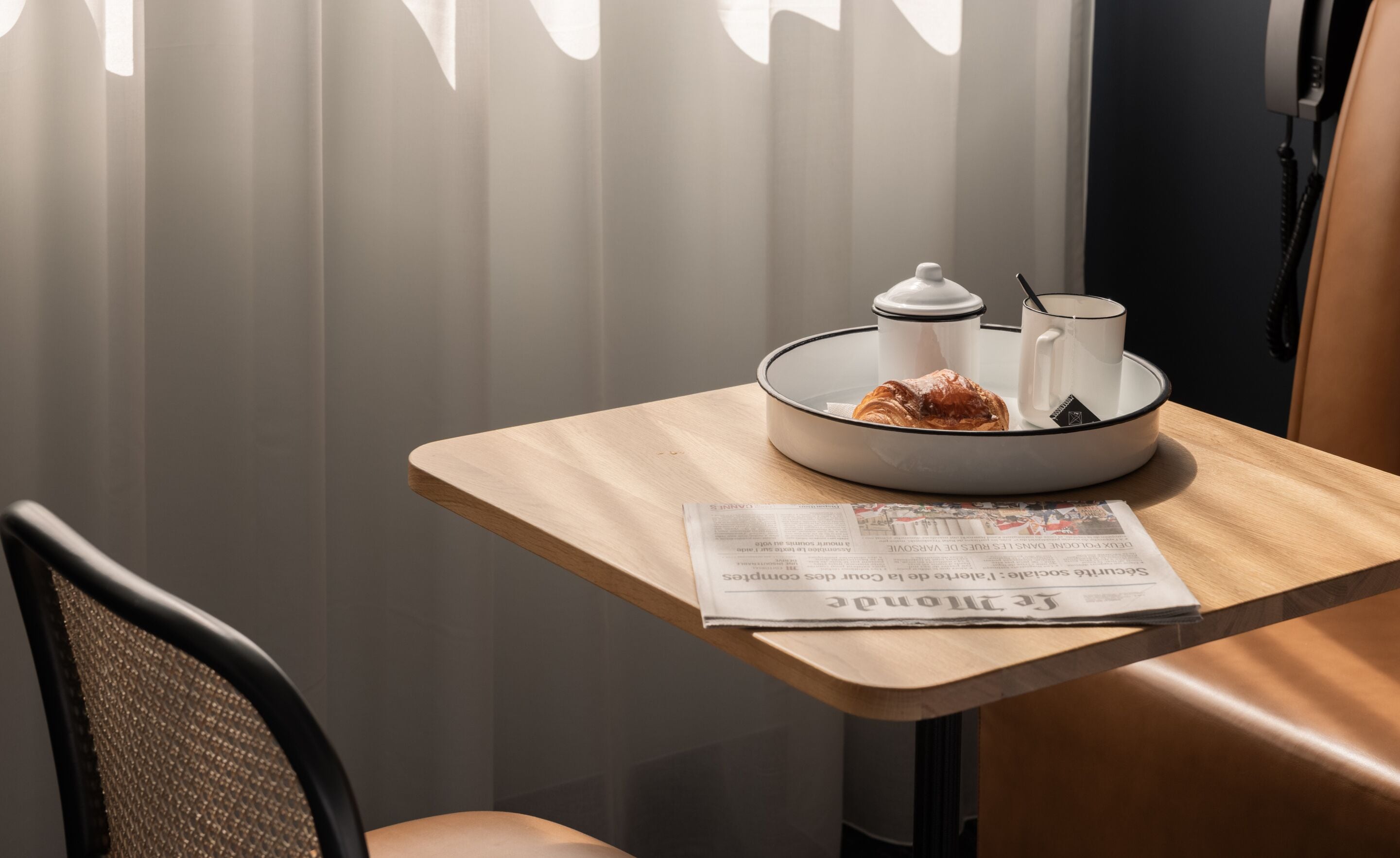 A breakfast setup with a croissant, a cup, and a teapot on a tray, a newspaper on the table, and a chair.