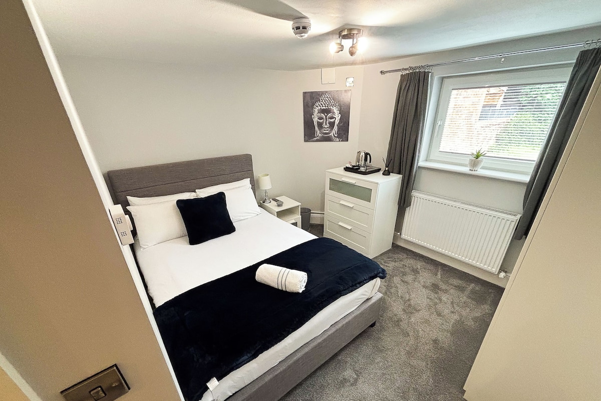 Ein ordentlich eingerichtetes Hotelzimmer mit Bett, Nachttisch, Lampe und einem Fenster mit Vorhängen.