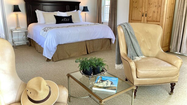 A hotel room with a large bed, a wooden wardrobe, a leather armchair, a glass coffee table with a book and a hat, and a small side table with a lamp. - Old Village Inn - King Room (Ogunquit)