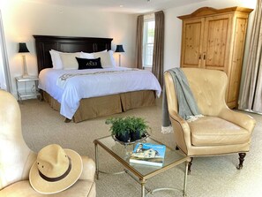 A hotel room with a large bed, a wooden wardrobe, a leather armchair, a glass coffee table with a book and a hat, and a small side table with a lamp. - Old Village Inn - King Room (Ogunquit)