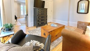 A living room with a wooden coffee table, a black leather sofa, a brown armchair, a black dresser with gold trim, and a flat-screen TV mounted on the wall. - Old Village Inn - King Room (Ogunquit)