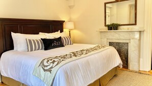 A neatly made bed with a headboard, a bedside lamp, and a fireplace with a mantel. - Old Village Inn - King Room (Ogunquit)