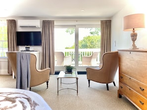 A hotel room with a bed, two armchairs, a small table, a dresser, a TV, and a balcony with wicker furniture. - Old Village Inn - King Room (Ogunquit)