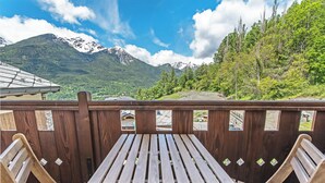 Una terraza de madera con una mesa de picnic y sillas, con vistas a un paisaje montañoso con picos nevados y abundante vegetación.