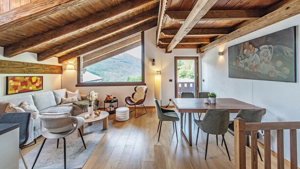 Un loft espacioso con techo alto, vigas de madera, una gran mesa de comedor con sillas, un sofá y una butaca con un jarrón de flores.