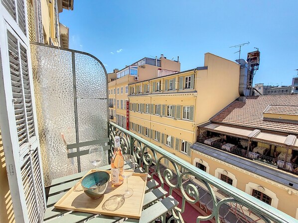 A balcony with a view of adjacent buildings, a table with a bowl and a bottle, and a railing.