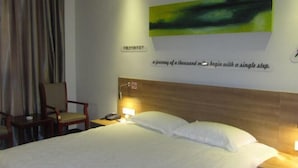 Quarto de hotel com cama, mesa de cabeceira com luminária, cadeira e parede com pintura abstrata verde e uma frase motivacional.