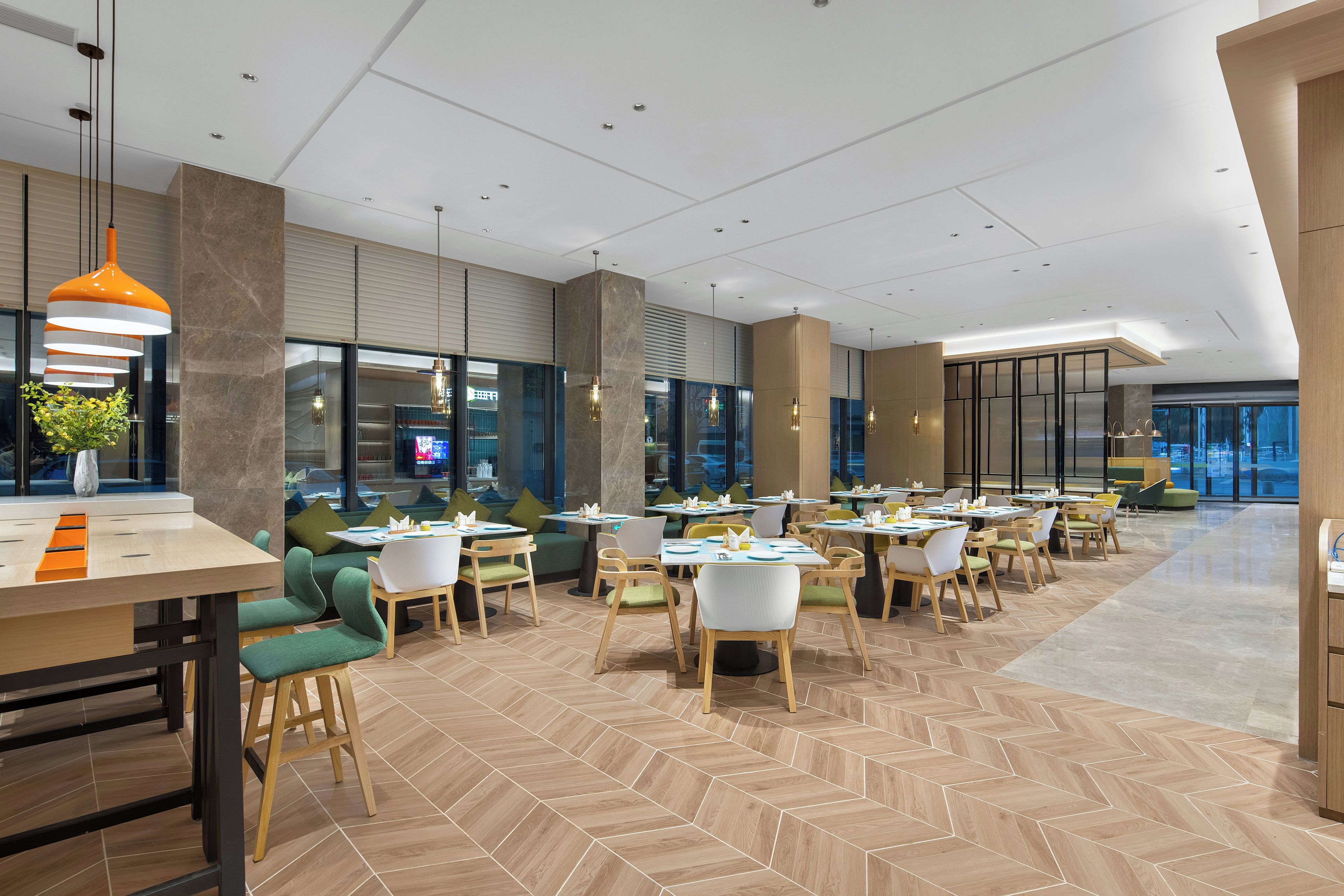 Un espace repas moderne doté de plusieurs tables et chaises, de grandes fenêtres et d’un sol à motifs en chevrons.