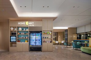 Un interior moderno con un quiosco 'HOME 2 MKT', un refrigerador lleno de bebidas y una zona de estar con sillas y una mesa.