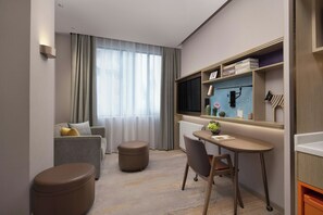 A compact hotel room with a sofa, a small table, a chair, a trash bin, a window with curtains, a television, and a shelf with books and decorative items. - Home2 Wuhan Hankou Railway Station (Wuhan)