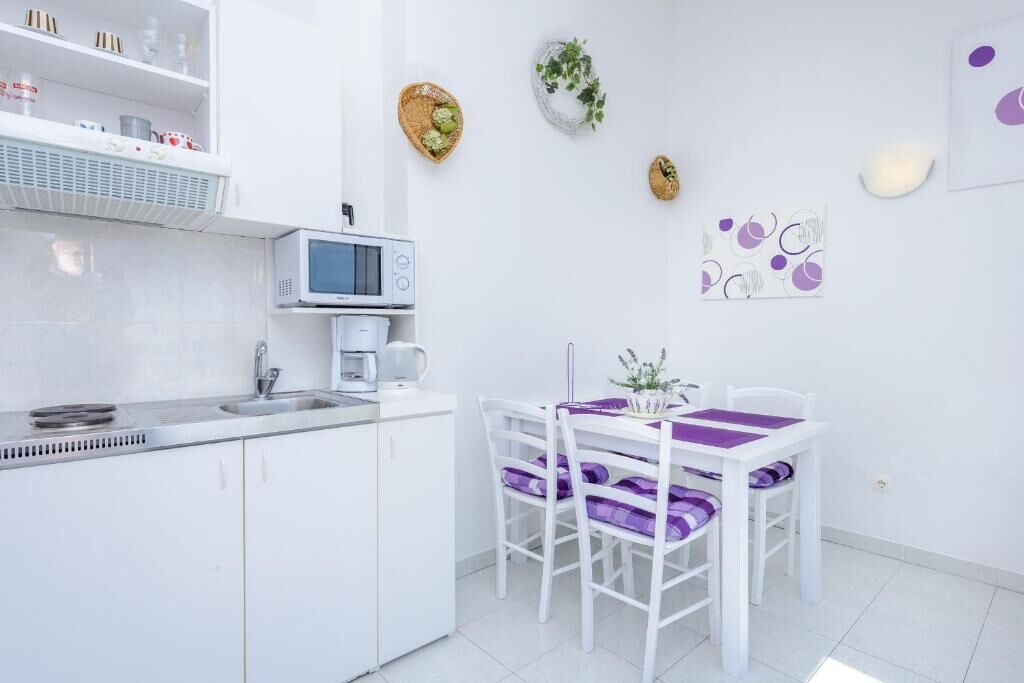 A modern kitchen with white cabinets, a microwave, and a coffee maker. There is a dining table with purple chairs and a small potted plant on it.