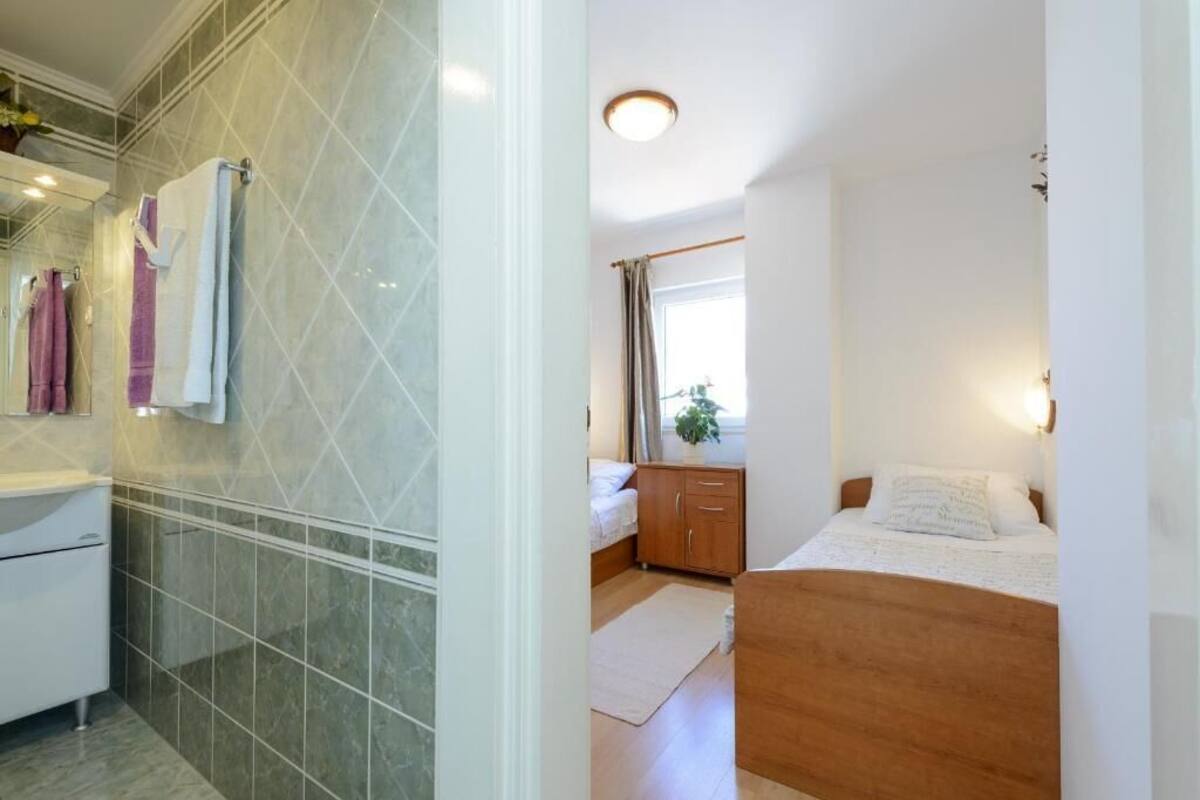 A bathroom with a shower, a sink, and a mirror. There is a bed with a wooden headboard and a window with curtains.