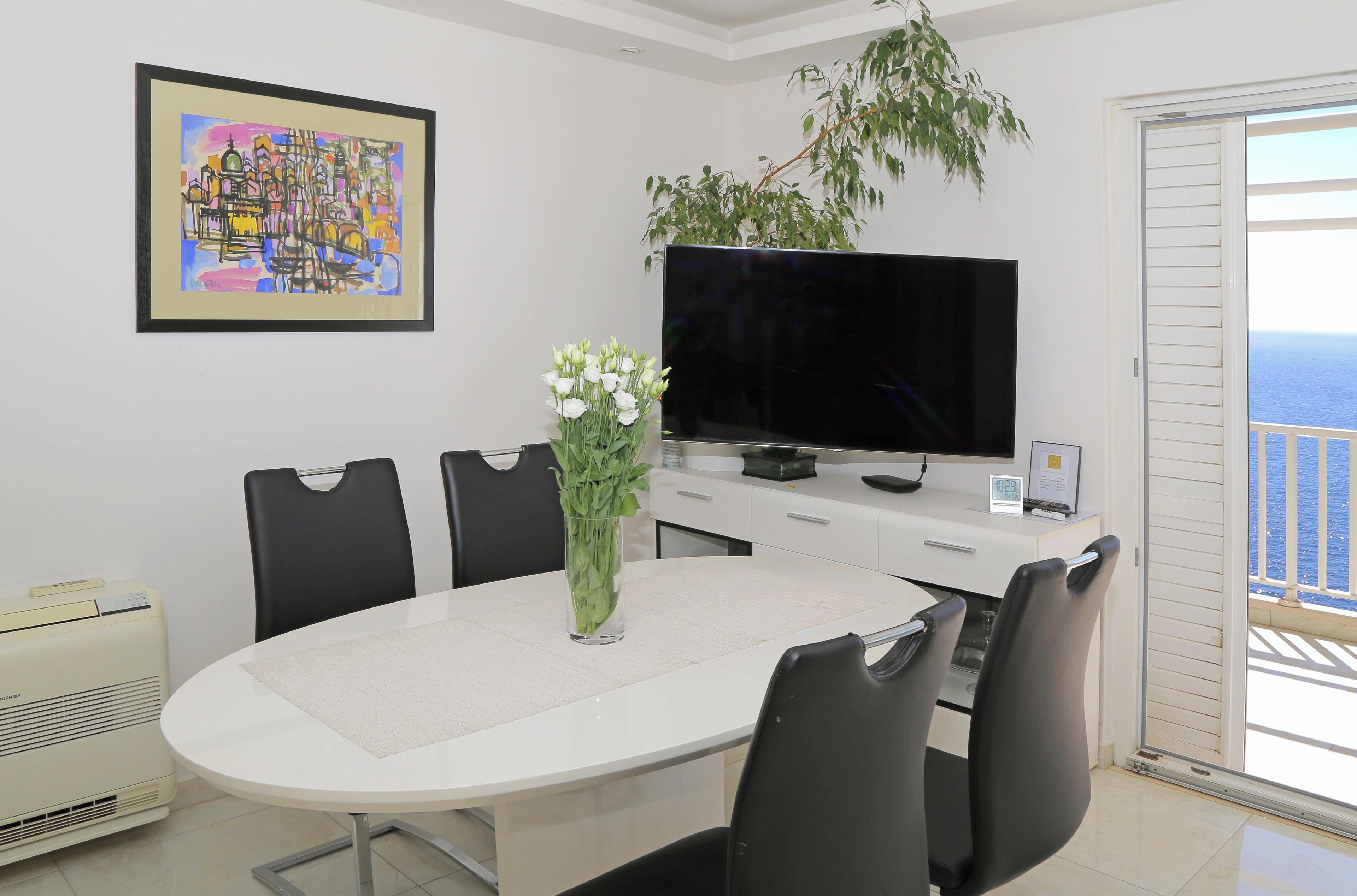 A modern dining area with a white table, black chairs, a flat-screen TV, a plant, and a painting on the wall.