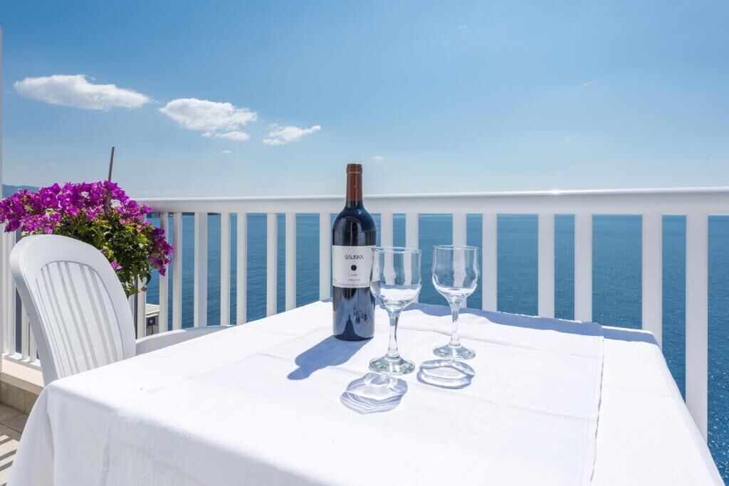 Een tafel met een fles wijn en twee glaasjes op een balkon met een witte reling en uitzicht op zee.