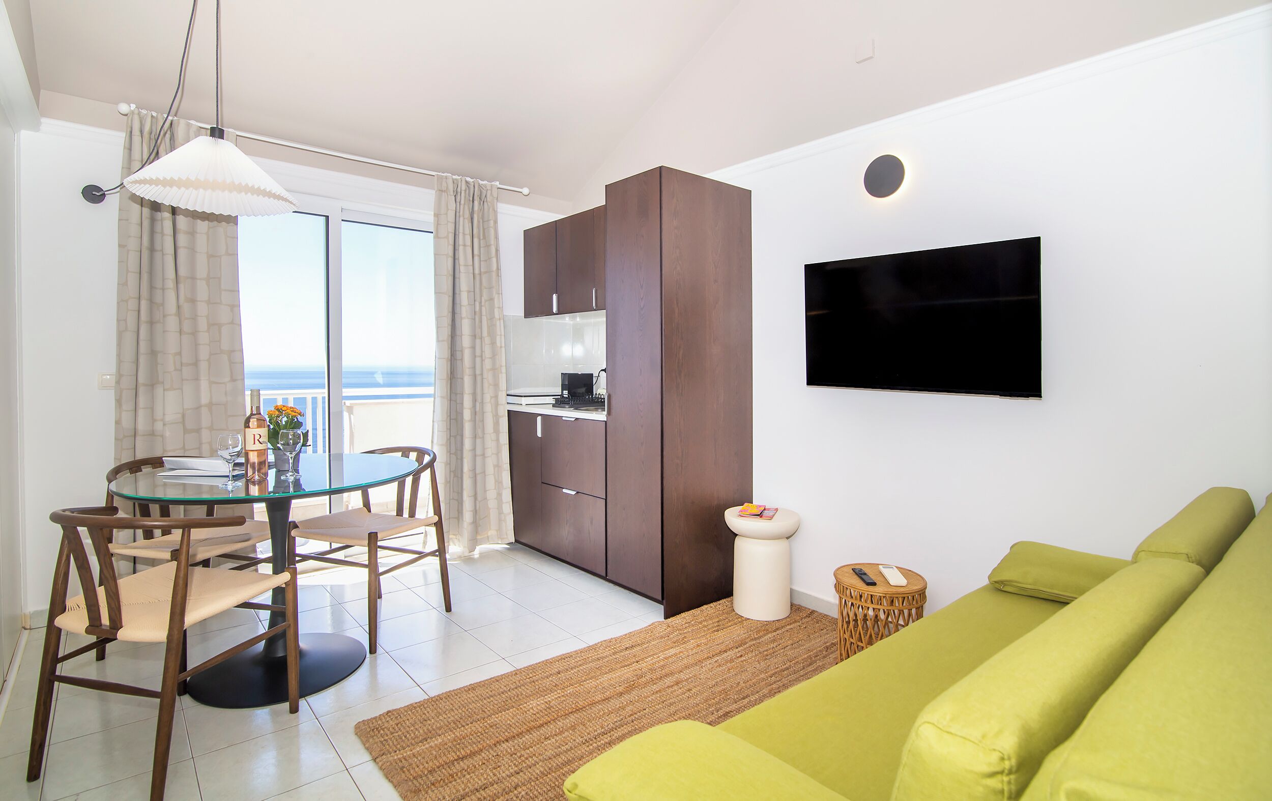 A modern living room with a green sofa, a dining table with chairs, a kitchen area, and a TV mounted on the wall.
