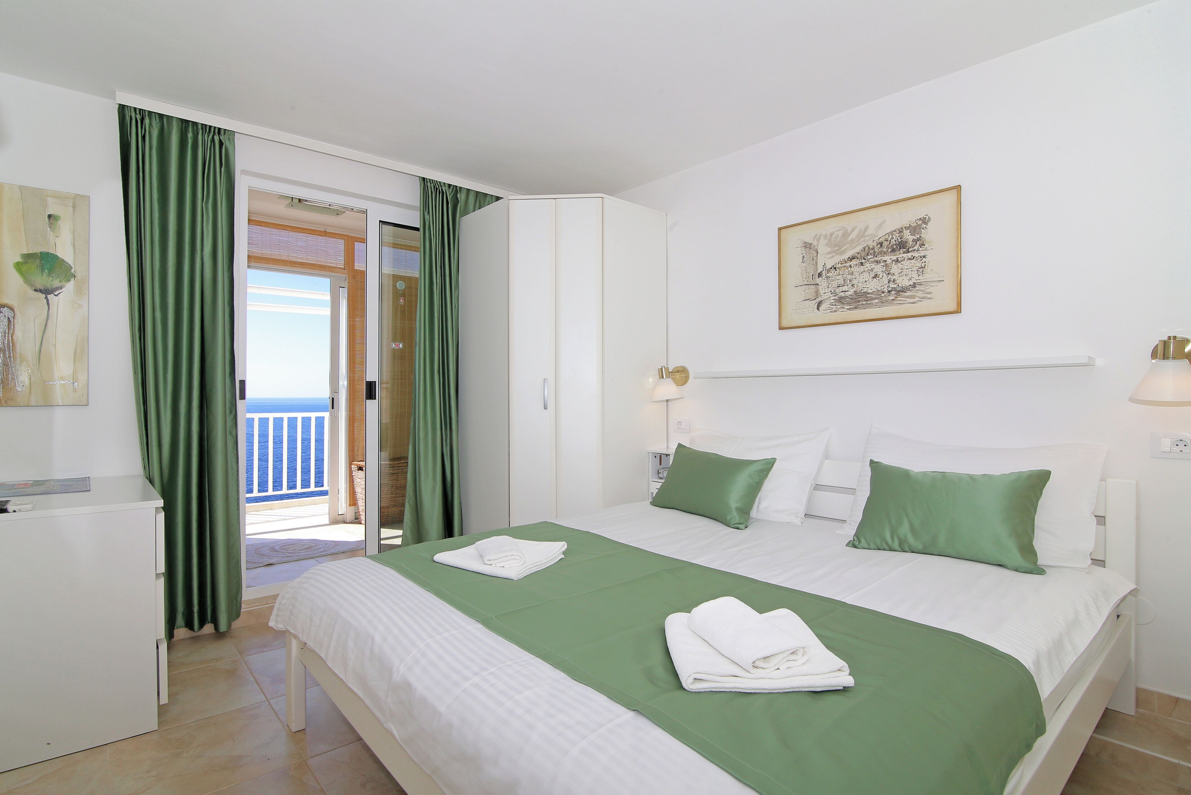 A hotel room with a bed, green curtains, a white desk, and a painting on the wall.