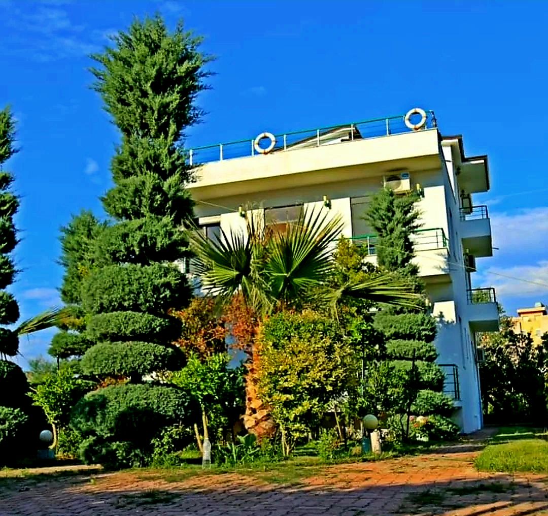 A modern building with balconies, surrounded by well-maintained greenery and a clear blue sky.
