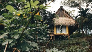 Une petite cabane au toit de chaume, peinte en jaune, entourée d’une végétation luxuriante et d’arbres.