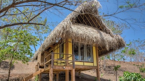 Une construction au toit de chaume, dotée d’une terrasse en bois et de fenêtres, entourée d’arbres et surplombant un ciel dégagé.