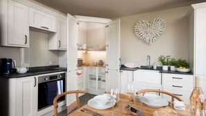 Une cuisine moderne avec des meubles blancs, un décor mural en forme de cœur et un coin repas aménagé pour deux personnes, avec une table en bois, des chaises et une miche de pain.