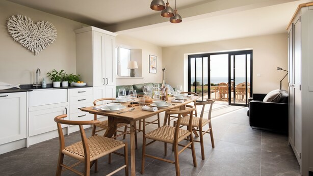 Une cuisine moderne avec un coin repas, comprenant une grande table dressée pour six personnes, des chaises en bois et une vue sur l’extérieur à travers la porte vitrée.