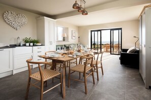 A modern kitchen with a dining area, featuring a large table set for six, wooden chairs, and a view through the glass door to the outdoors.